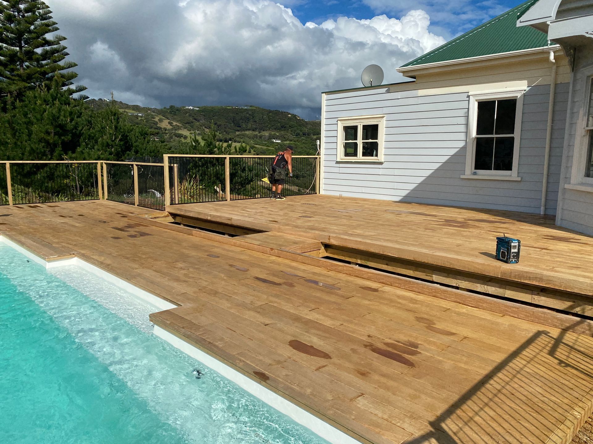 Wooden deck beside a turquoise pool, with a house under construction and mountains in the background