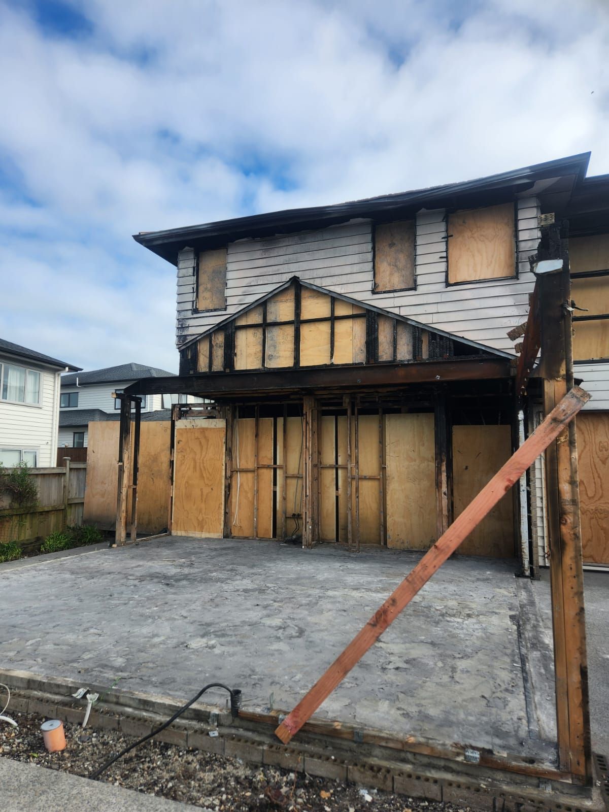 Boarded-up two-story house with damaged facade and leaning wooden support beam on a vacant lot