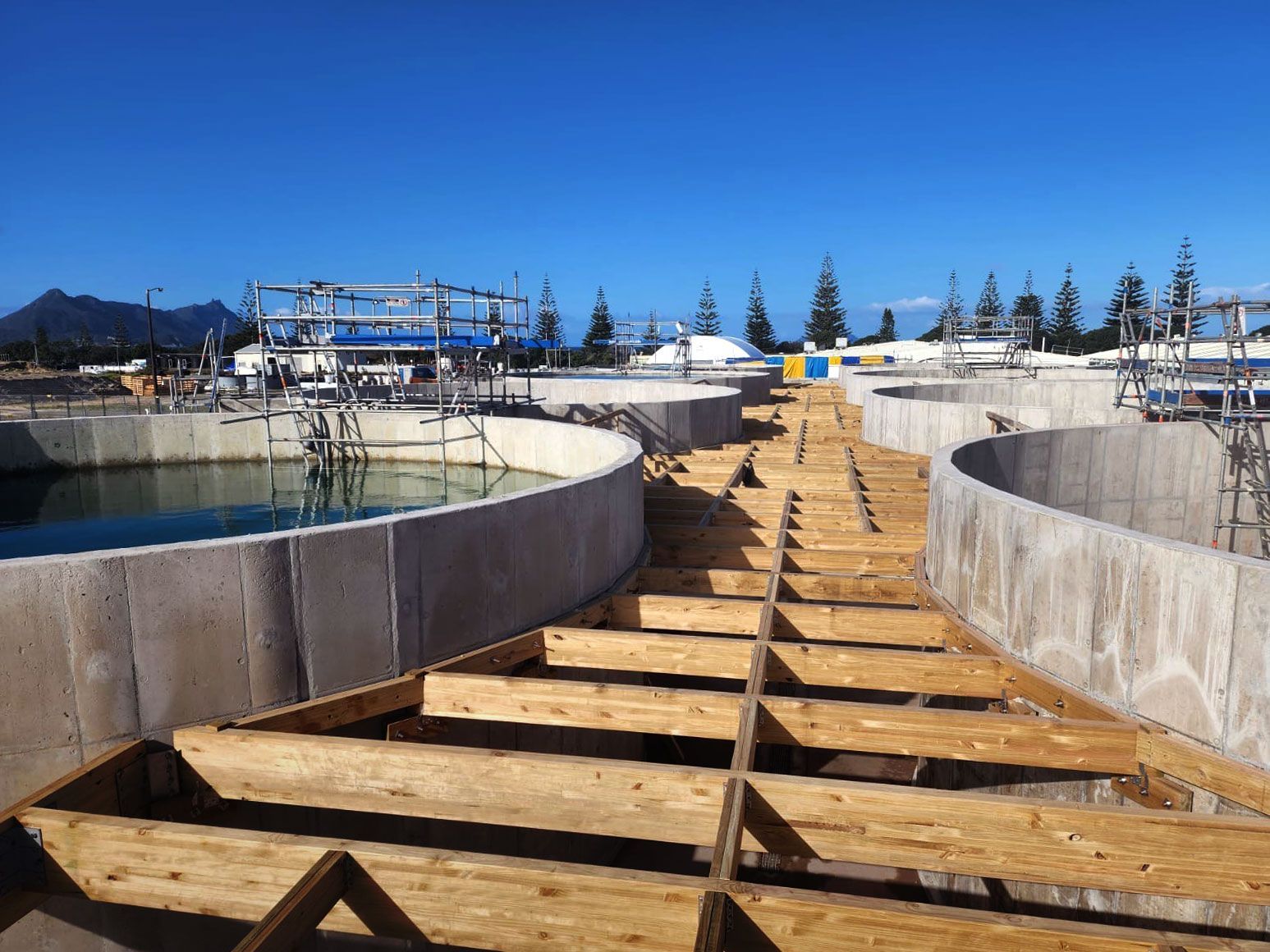Wooden boardwalk between concrete tanks at a marina under a clear blue sky