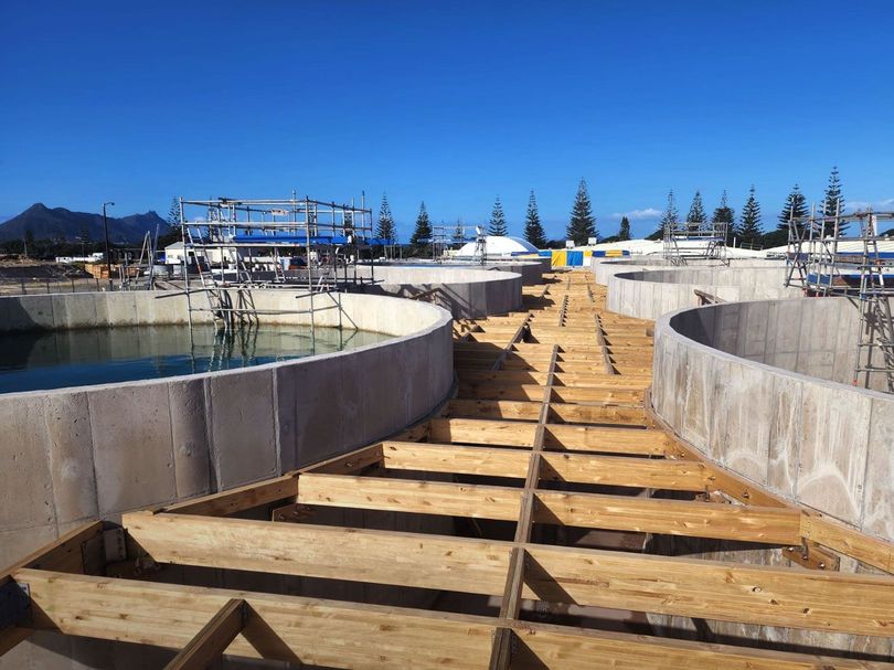 Wooden walkway between large concrete basins at a sunny construction site under a clear blue sky