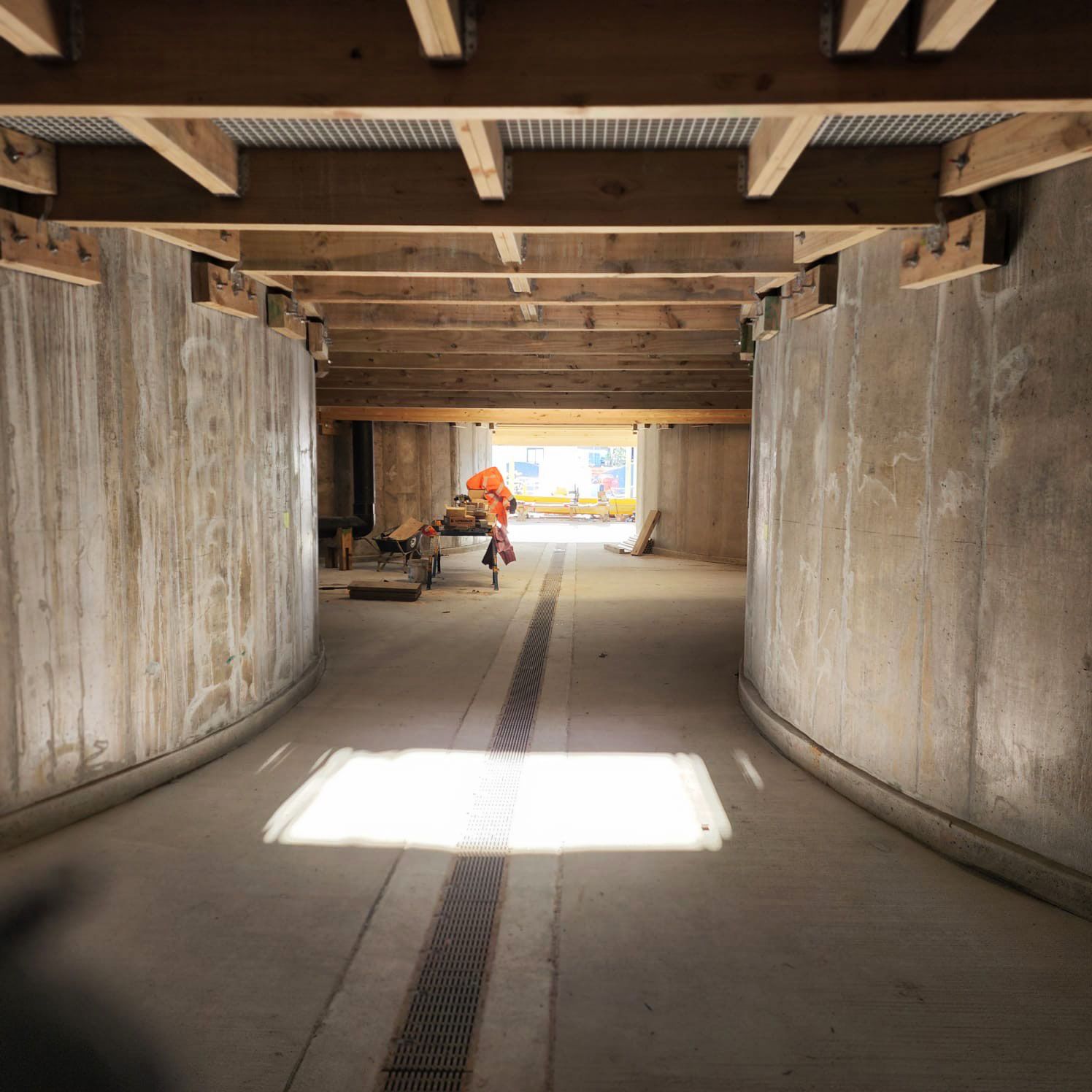 Inside a concrete tunnel, a worker crouches near the far end under wooden beams with bright light ahead.