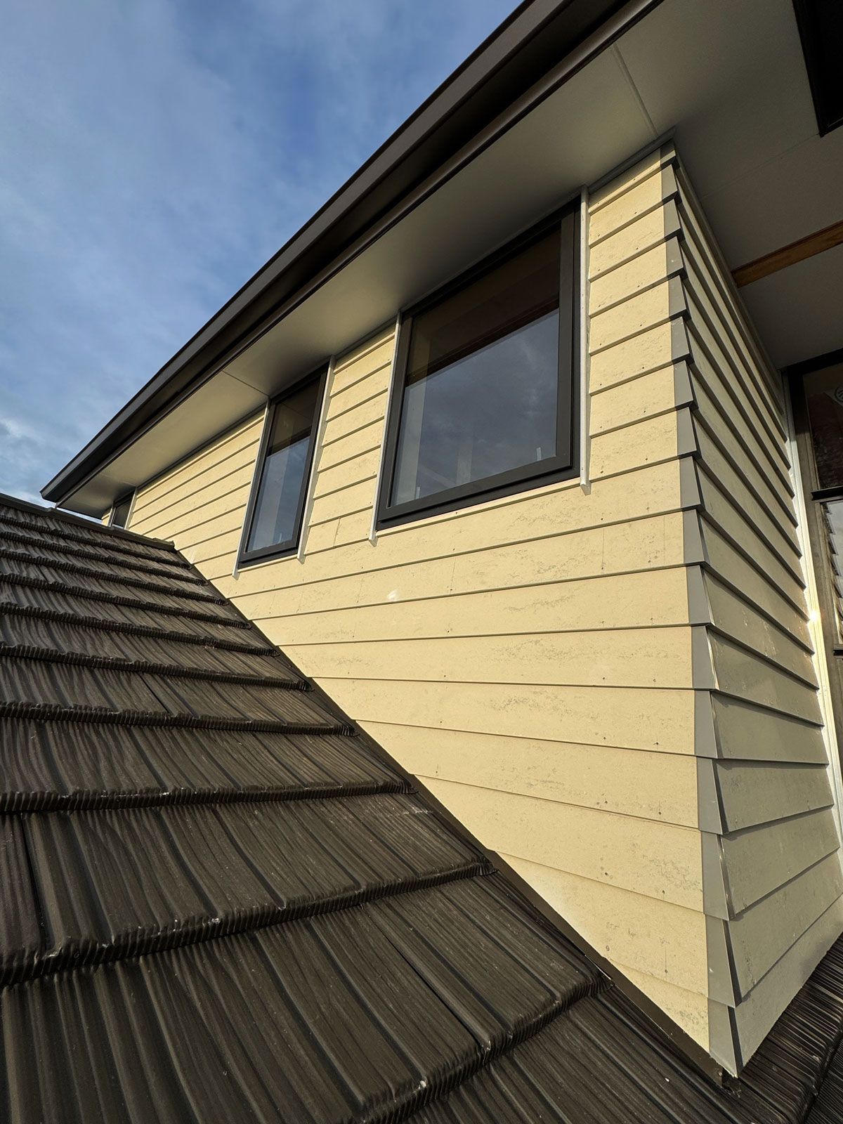 Roof and beige house siding with two windows under a blue sky
