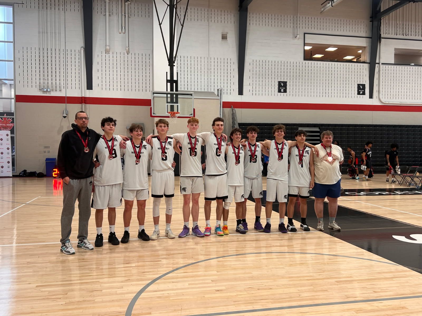 Basketball team posing for a photo in a gym. Players wearing black and white uniforms, coaches standing in the background.