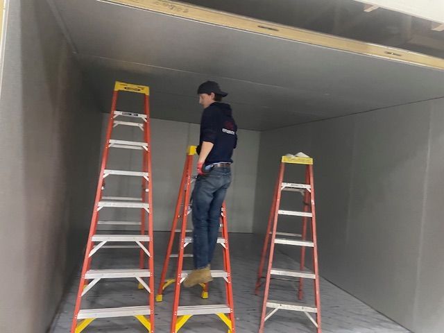 Worker on a ladder inside a large empty room, inspecting the ceiling with two other ladders nearby.