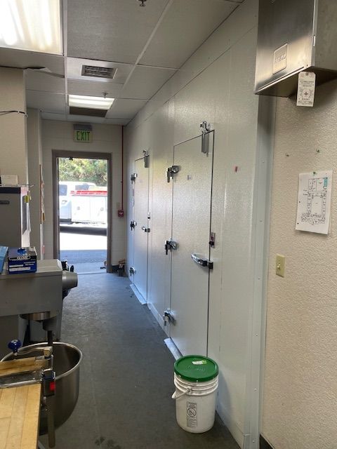 Narrow commercial kitchen hallway with hanging tools, white walls, and an open door to daylight.
