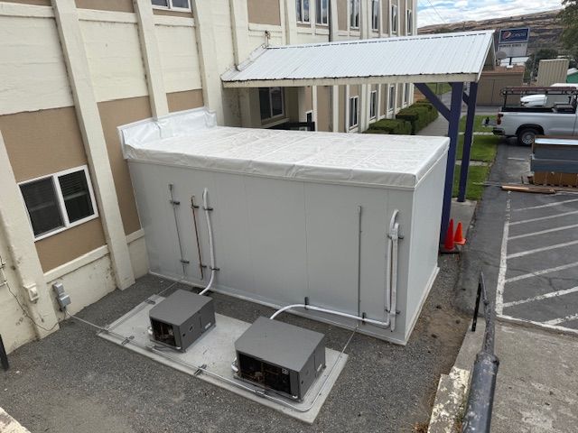 Two gray HVAC units beside a white utility enclosure in a parking lot under a covered walkway.