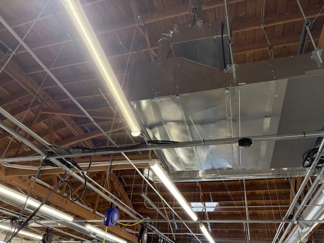 Industrial ceiling with exposed wooden beams, silver ductwork, and fluorescent lights inside a warehouse.