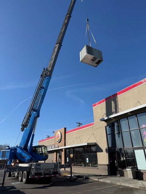 Crane lifting a large HVAC unit beside a storefront under a clear blue sky