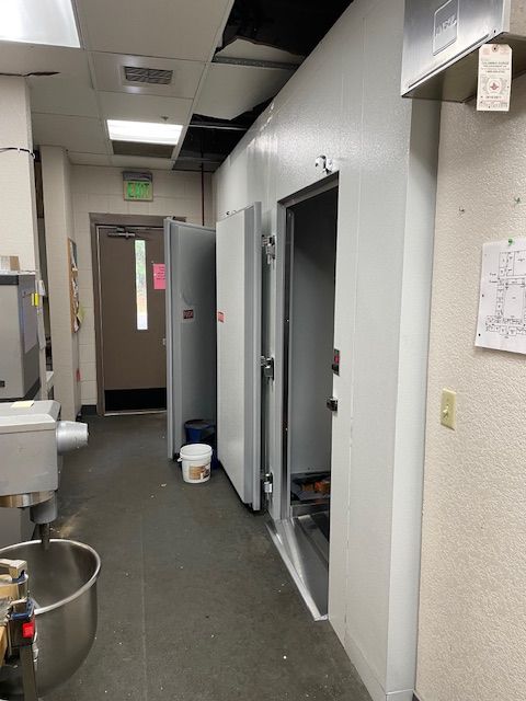 Narrow institutional hallway with open utility doors, gray walls, and a bucket on the floor