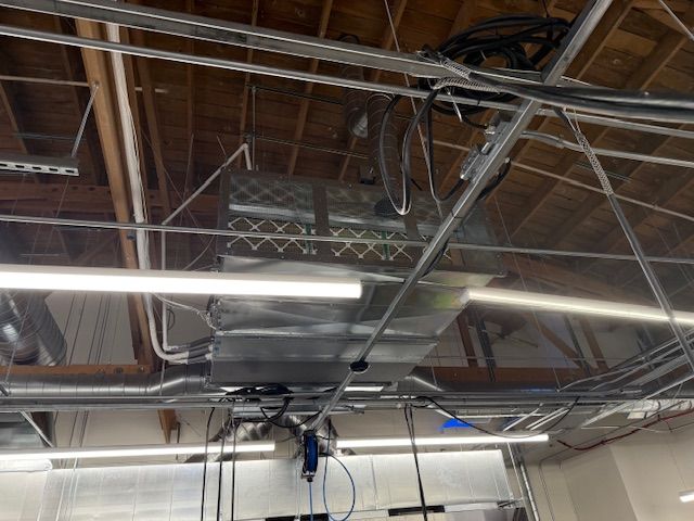 Industrial ceiling with exposed ducts, metal beams, fluorescent lights, and hanging cables.