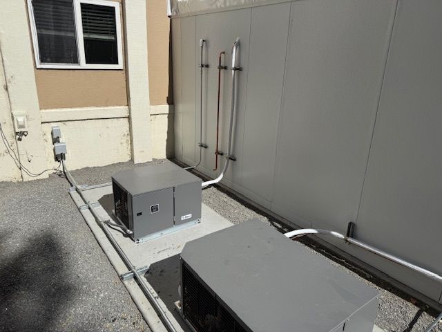 Outdoor HVAC units beside a building wall, with gray utility equipment and piping on a concrete pad.