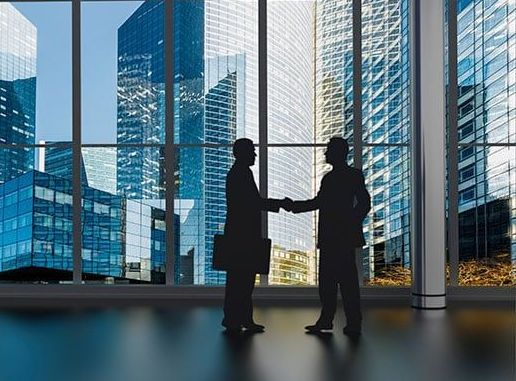 Two men are shaking hands in front of a window with a city in the background.