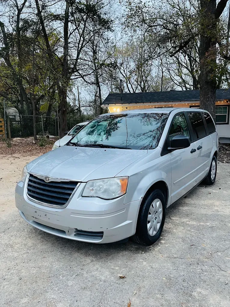 Silver minivan parked on a dirt road beside trees and a small building.