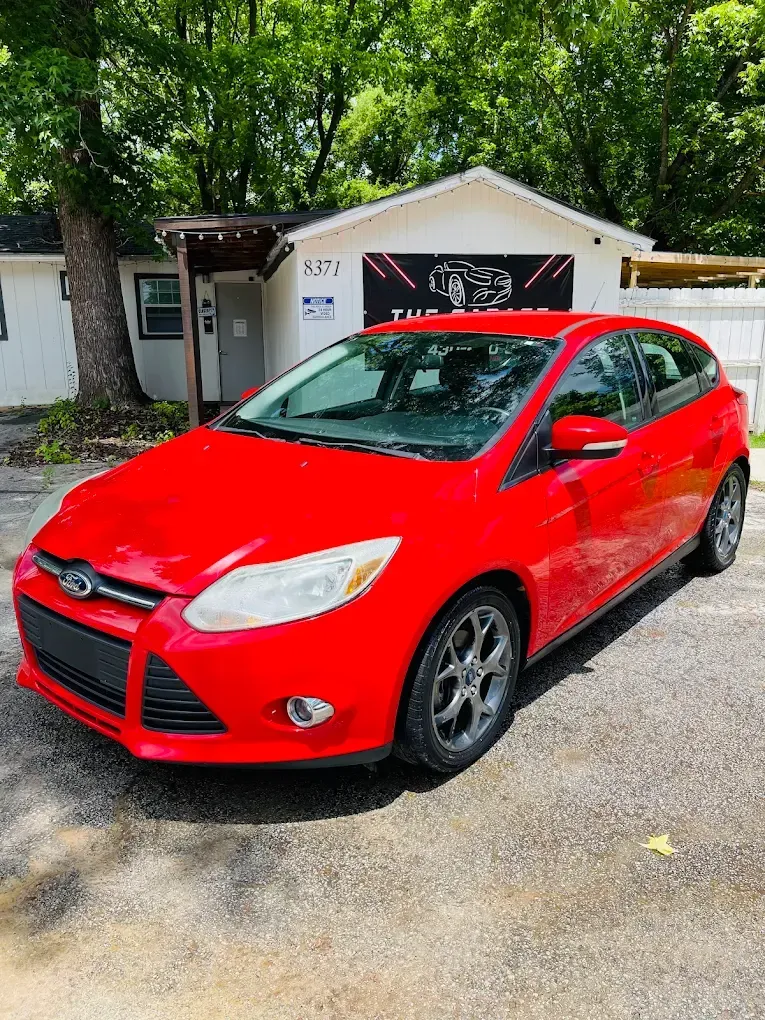 Red compact car parked in a driveway in front of a white garage surrounded by trees.