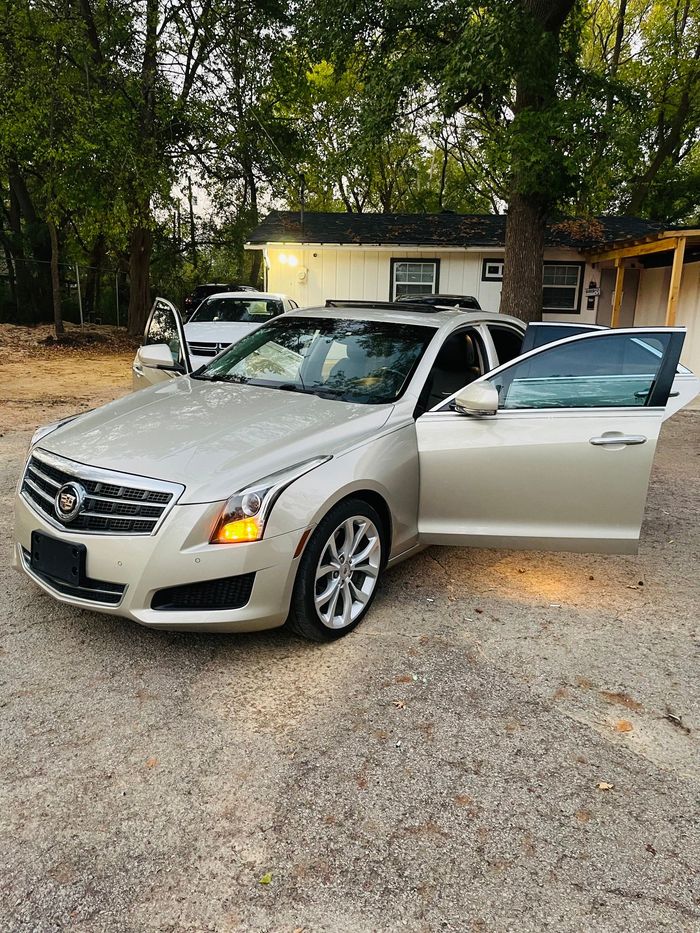 Silver sedan parked on gravel with the driver’s door open in front of a small house.