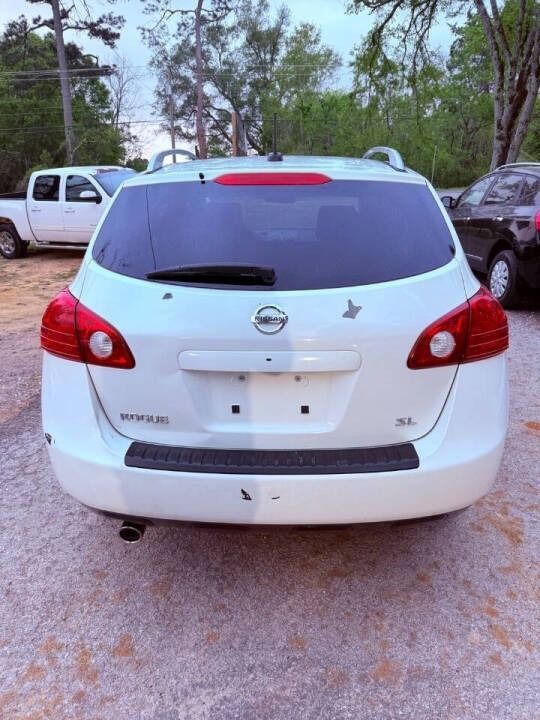 Rear view of a white Nissan sedan parked on a gravel lot with trees and other vehicles nearby.
