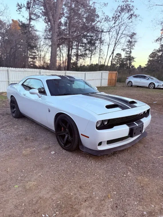White Dodge Challenger parked on gravel, black wheels and hood vents, with trees and a white van in the background