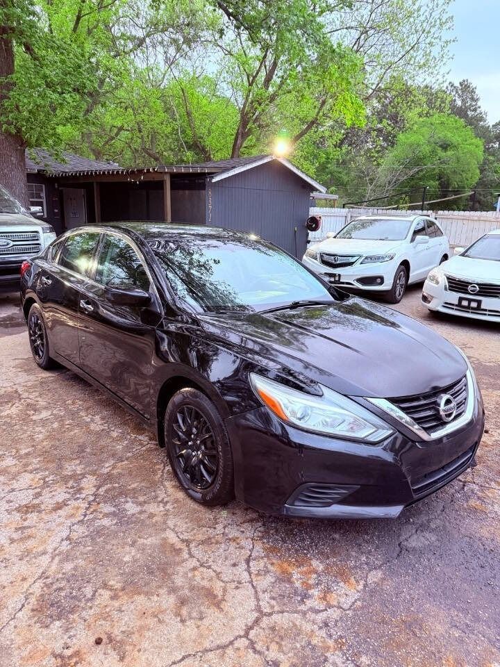 Black sedan parked in a driveway beside a house, with other cars and trees in the background.