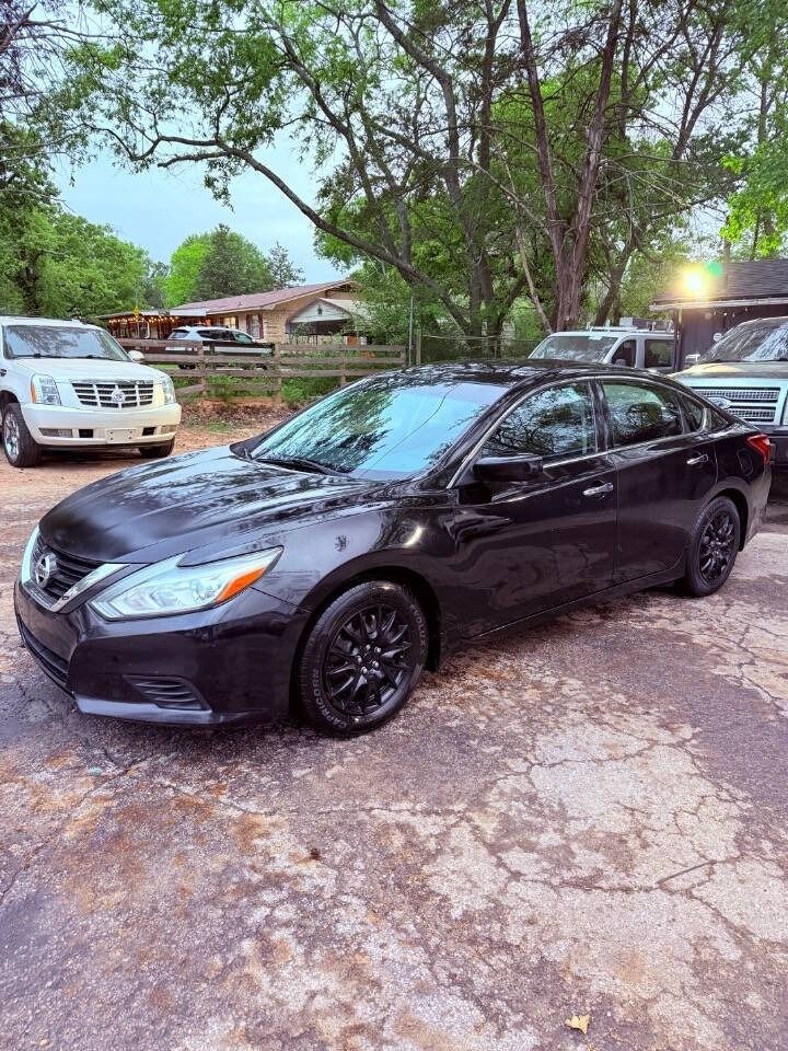 Black sedan parked on a dirt road under trees, with a white SUV and houses in the background.