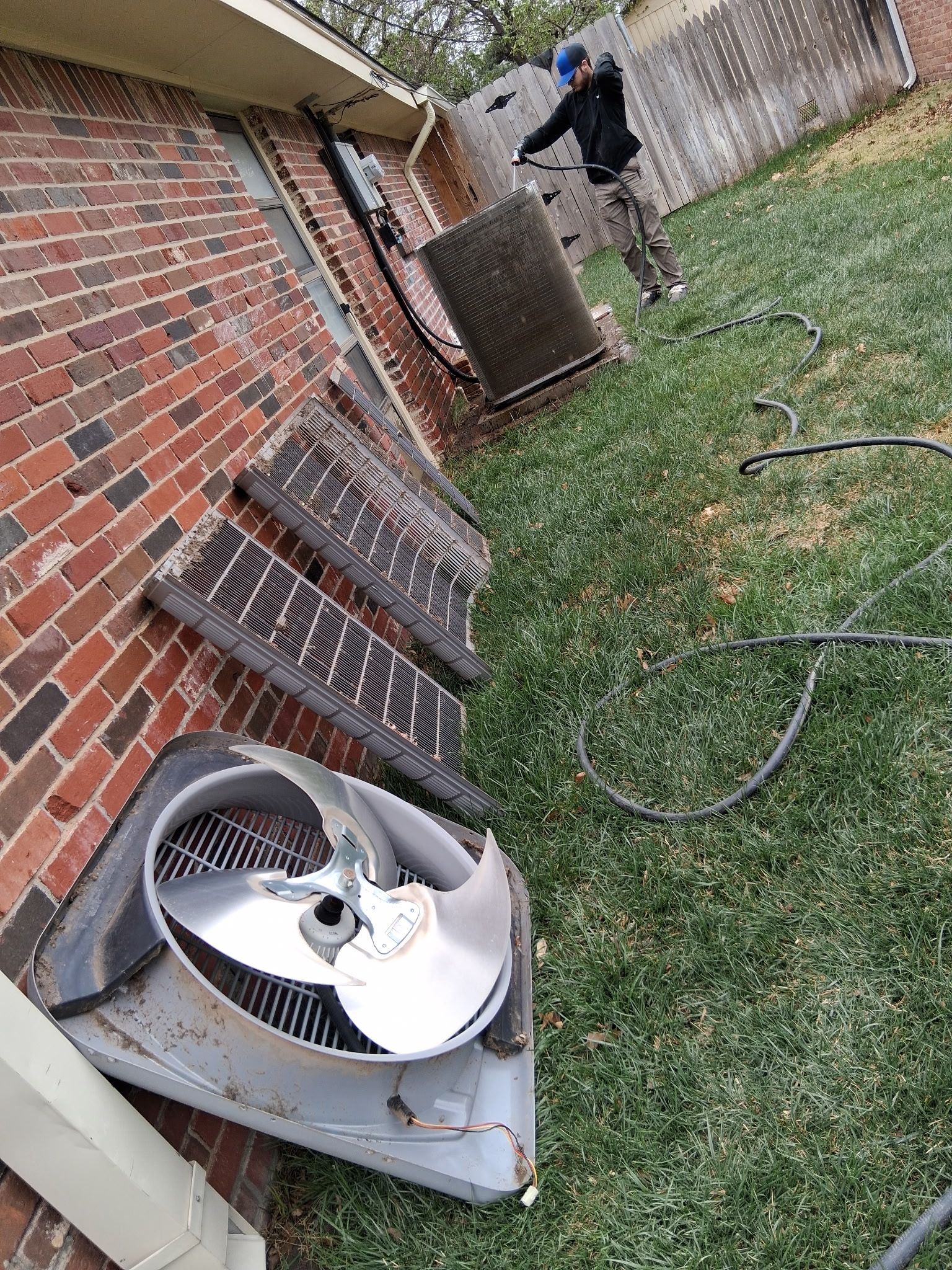 A person in a yard cleaning detached air conditioning unit panels leaning against a brick house wall.
