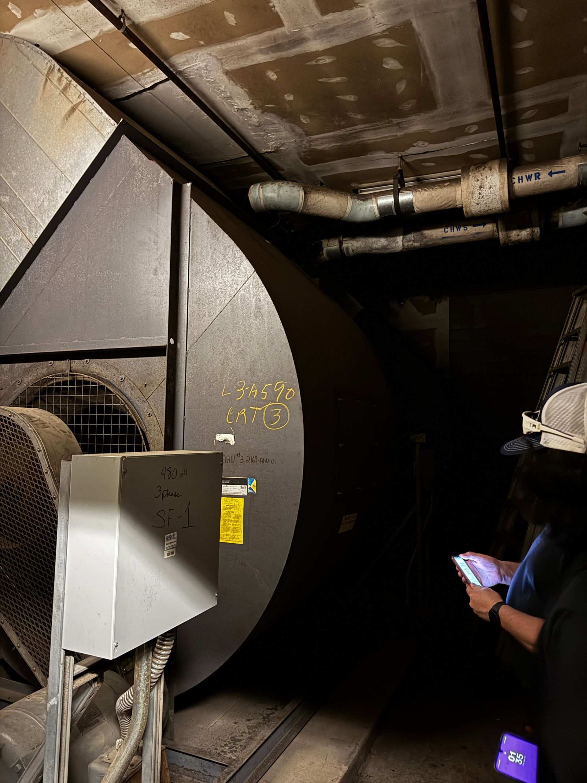 A technician in a gray hoodie works on an outdoor HVAC unit using a digital manifold gauge set and a power drill.