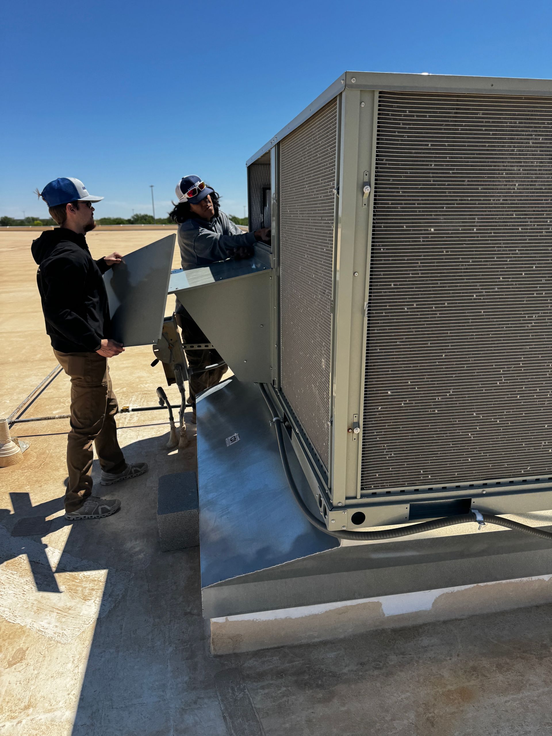 Two technicians work on an HVAC unit in an attic, one checking a handheld device and the other wearing a respirator mask.
