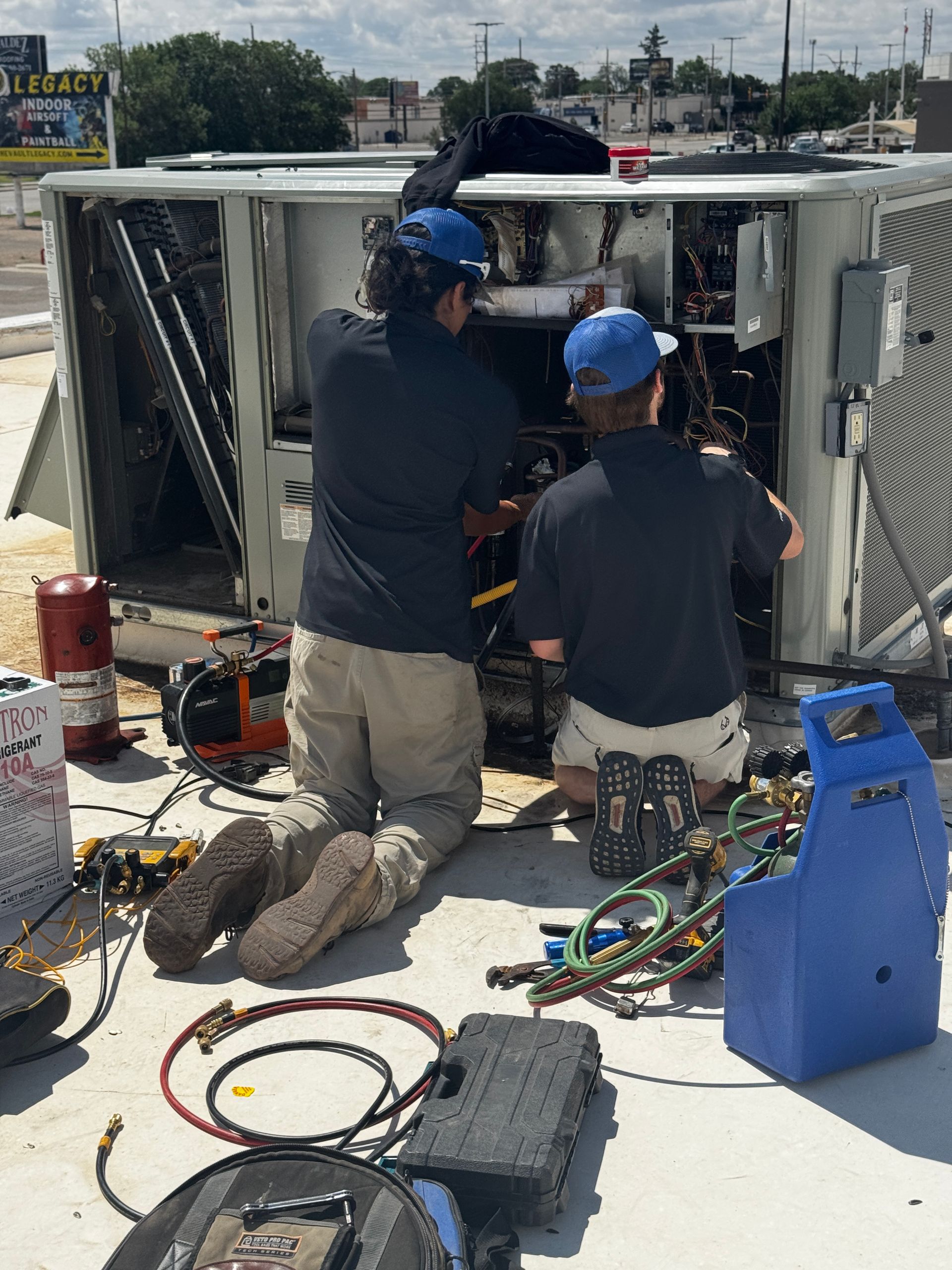 Two people examine the open electrical panel of a furnace in a dark space, one pointing to a wiring diagram.