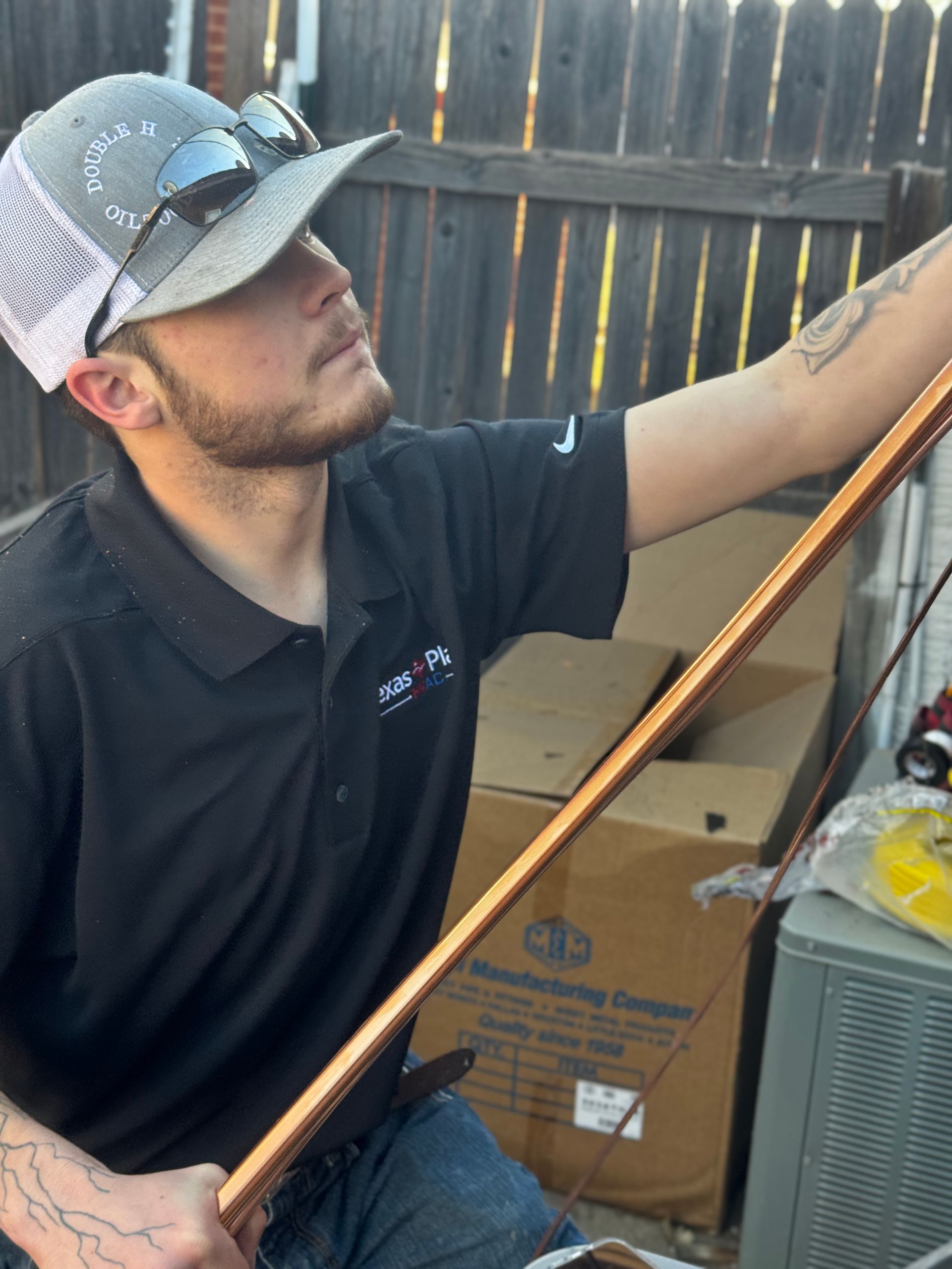 A person in a black polo and cap holds a length of copper pipe, working in an outdoor yard with a cardboard box nearby.