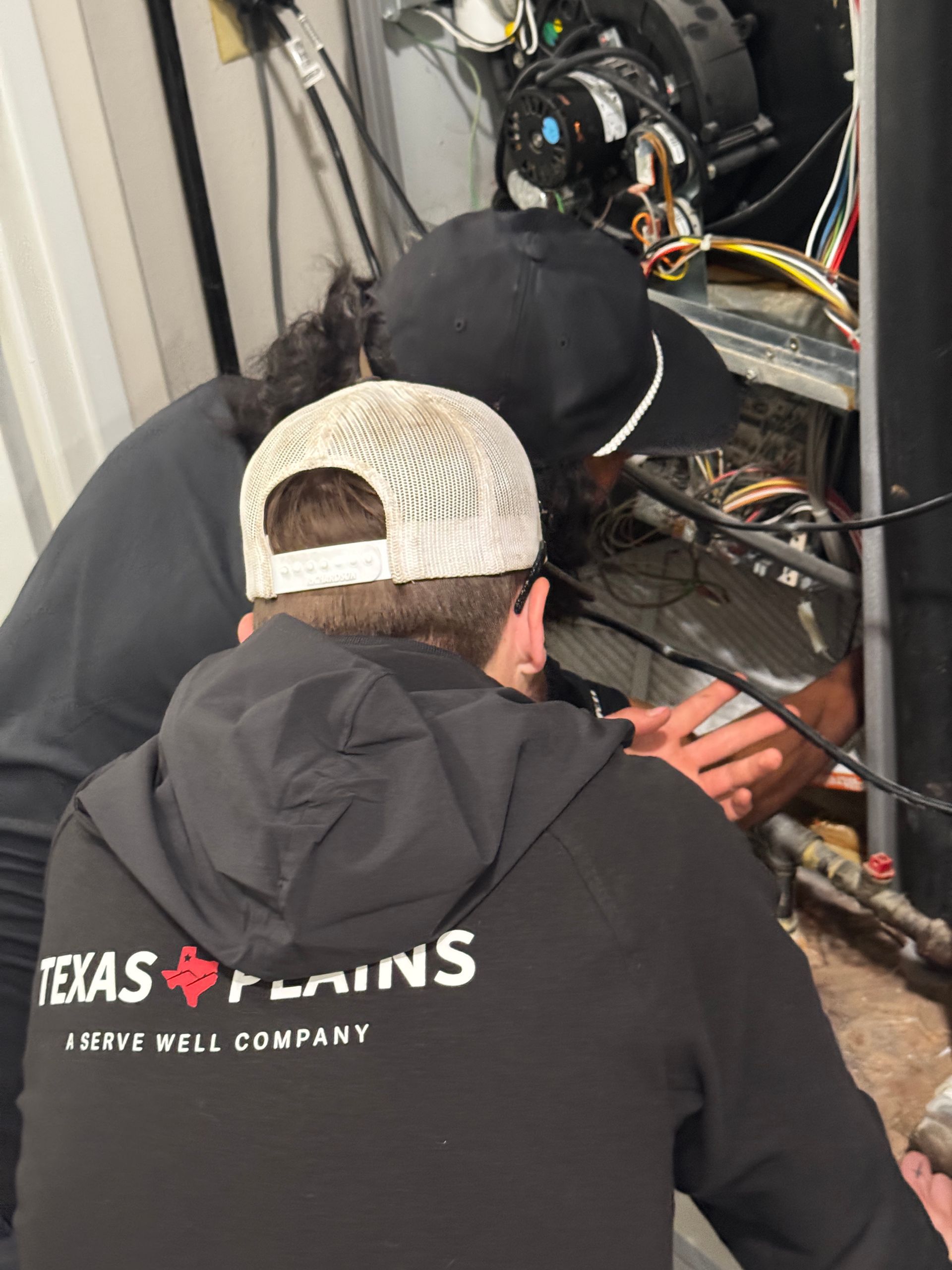 Two workers in dark uniforms perform maintenance on an indoor furnace unit.