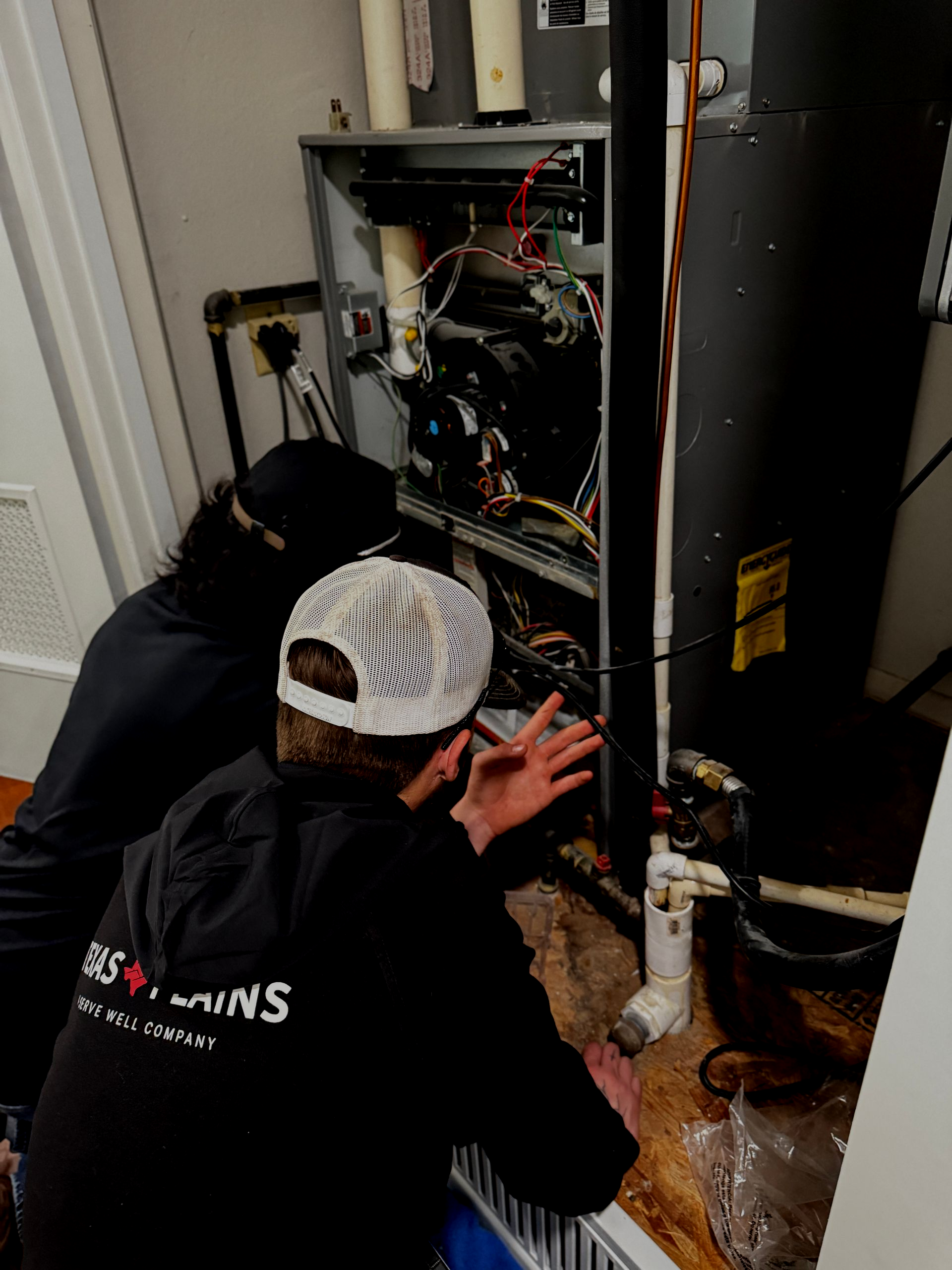 Two technicians in uniforms and hats inspect the internal components of a furnace unit in a residential utility room.