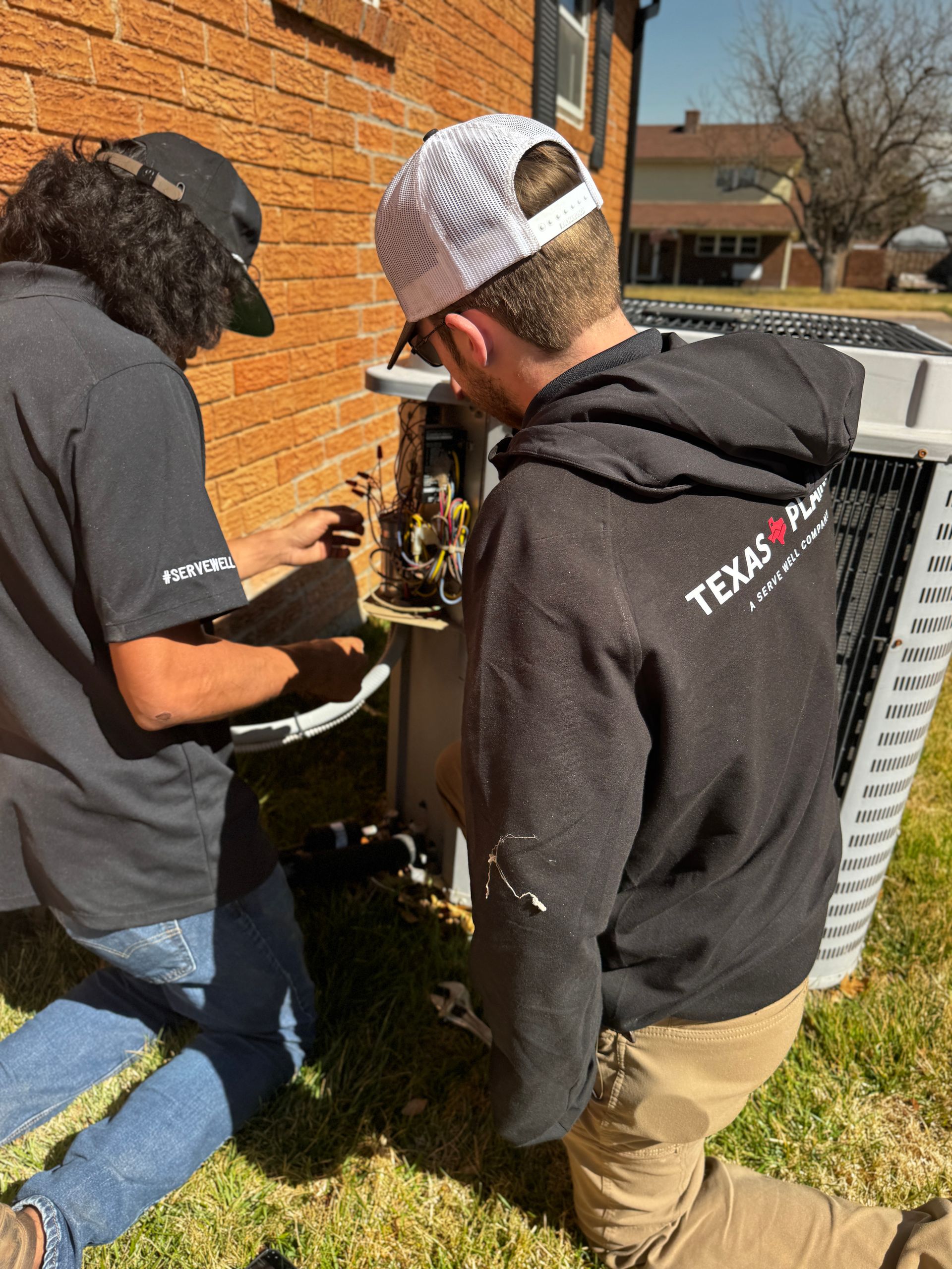 Two workers in uniforms repair the exposed electrical components of an outdoor residential air conditioning unit.