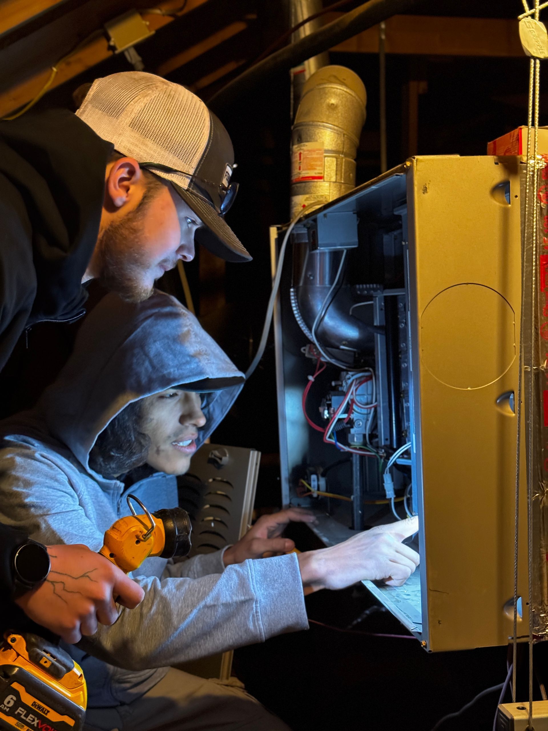 Two people examine the open interior of a furnace in a dimly lit attic while holding a flashlight.