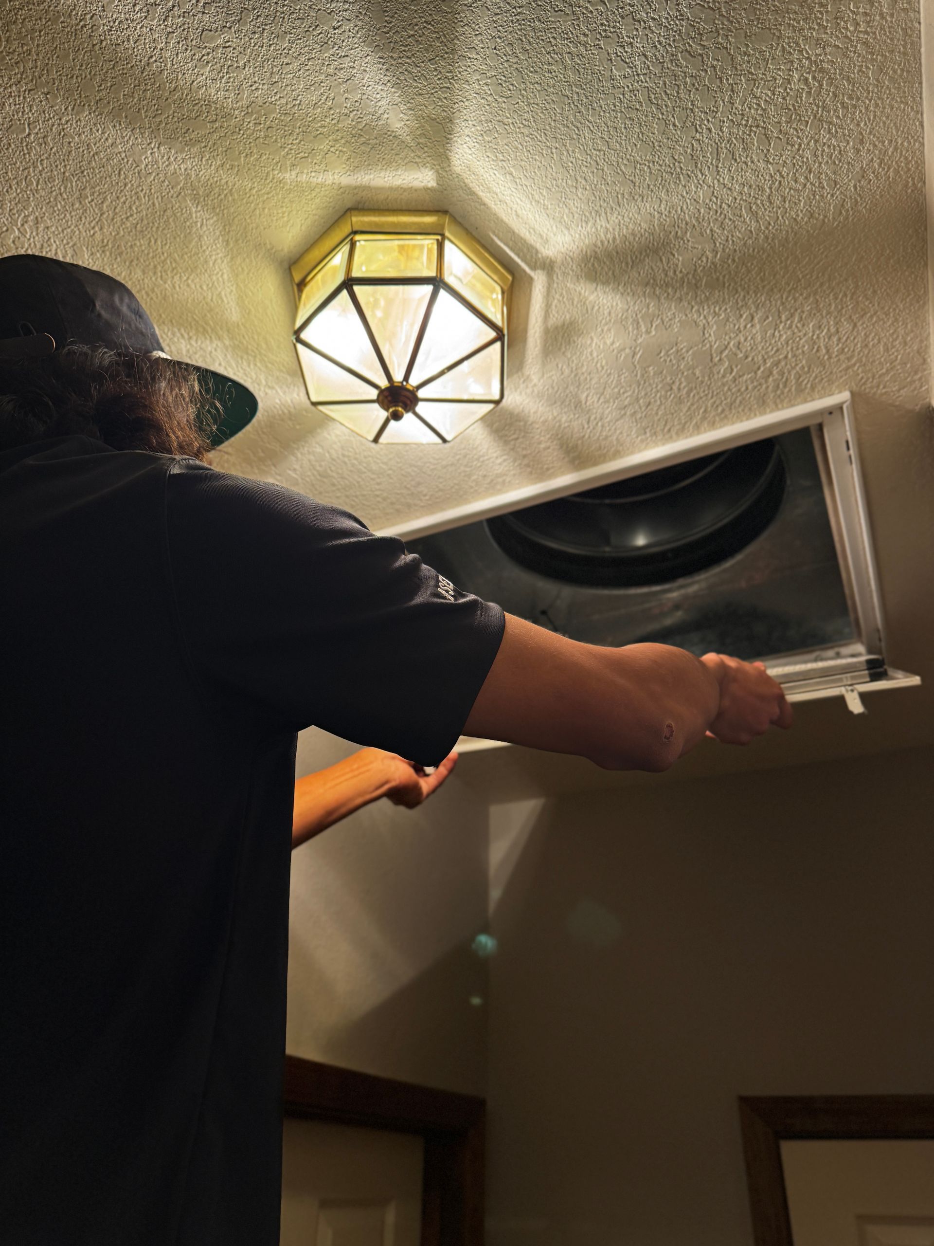 A person in a dark shirt stands on a ladder, opening a white ceiling HVAC vent cover near a geometric light fixture.