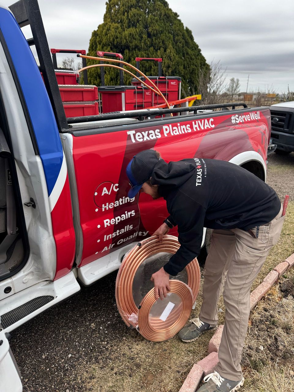 A technician in a gray hoodie works on an outdoor HVAC unit using a digital manifold gauge set and a power drill.