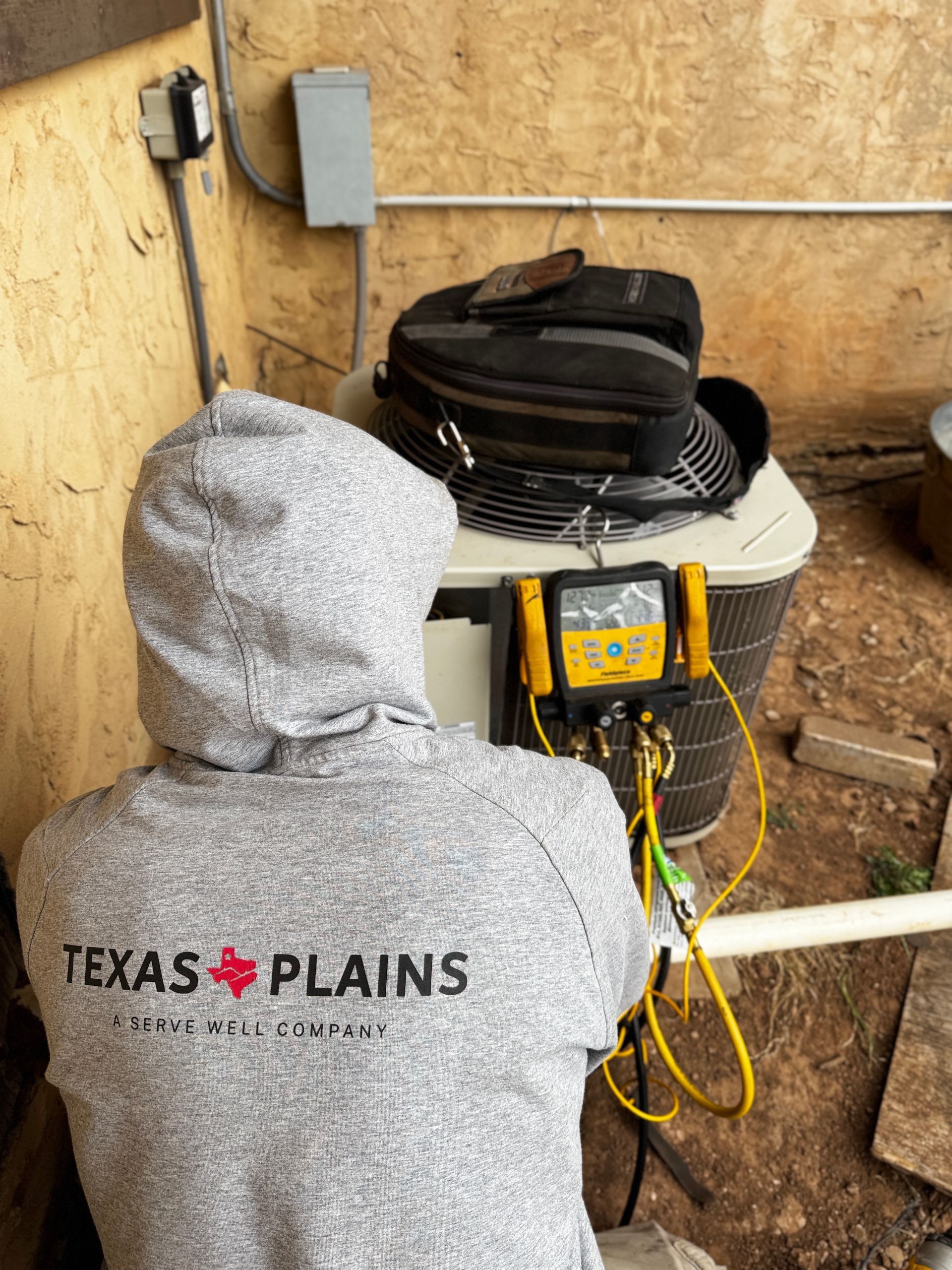 A technician in a gray Texas Plains hoodie repairs an outdoor HVAC unit using a digital manifold gauge.