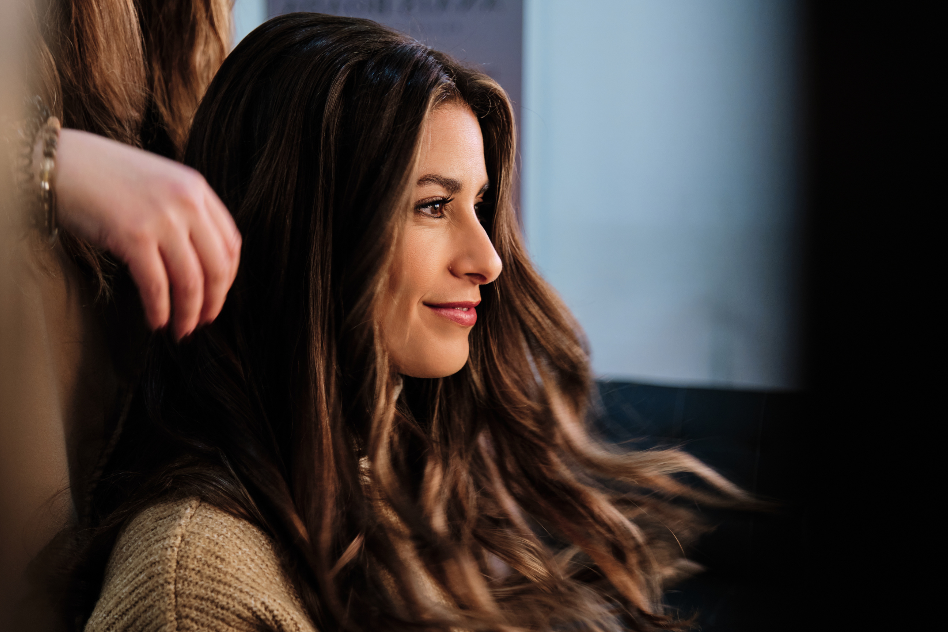 Woman with long brown hair smiling in profile indoors, with a blurred hand near her shoulder