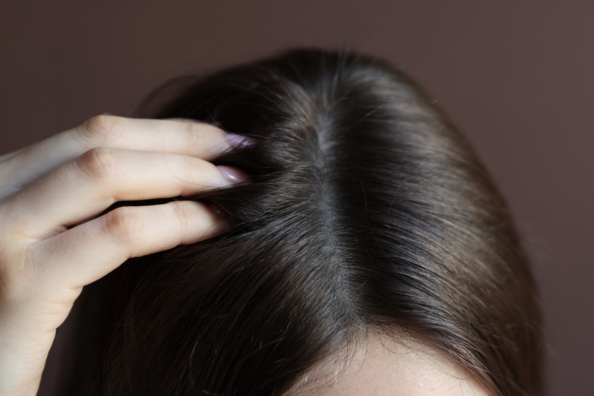 Hand parting dark brown hair at the scalp, showing a close-up of the top of the head