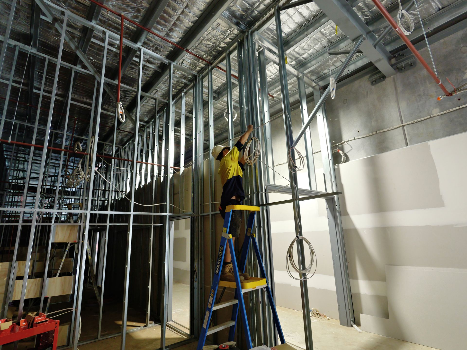 Construction Worker on a Ladder Installing Metal Studs for a Wall — Rob Ward Electrical Services in Kingscliff, NSW