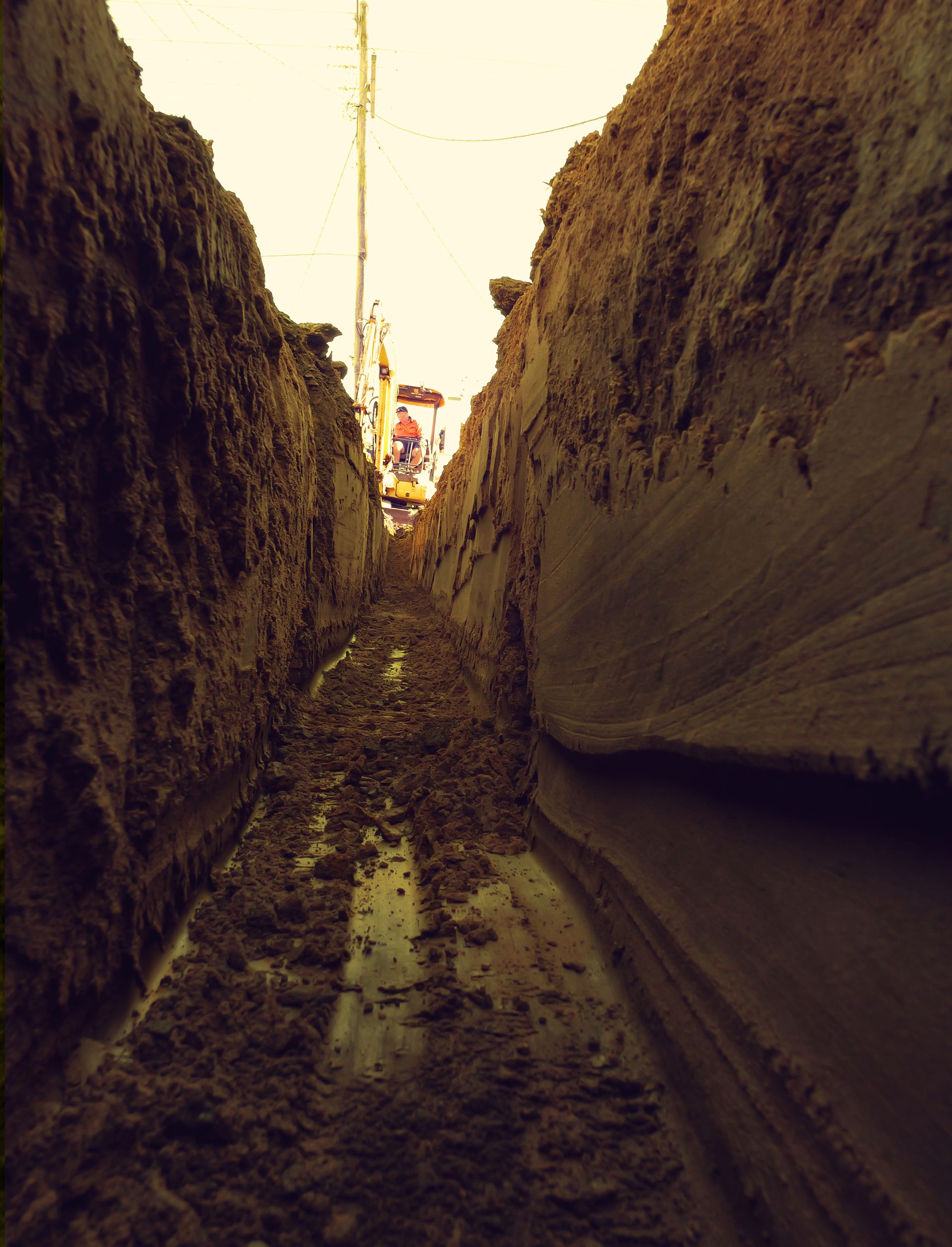 Narrow Muddy Trench With Backhoe in the Distance — Rob Ward Electrical Services in Alstonville, NSW