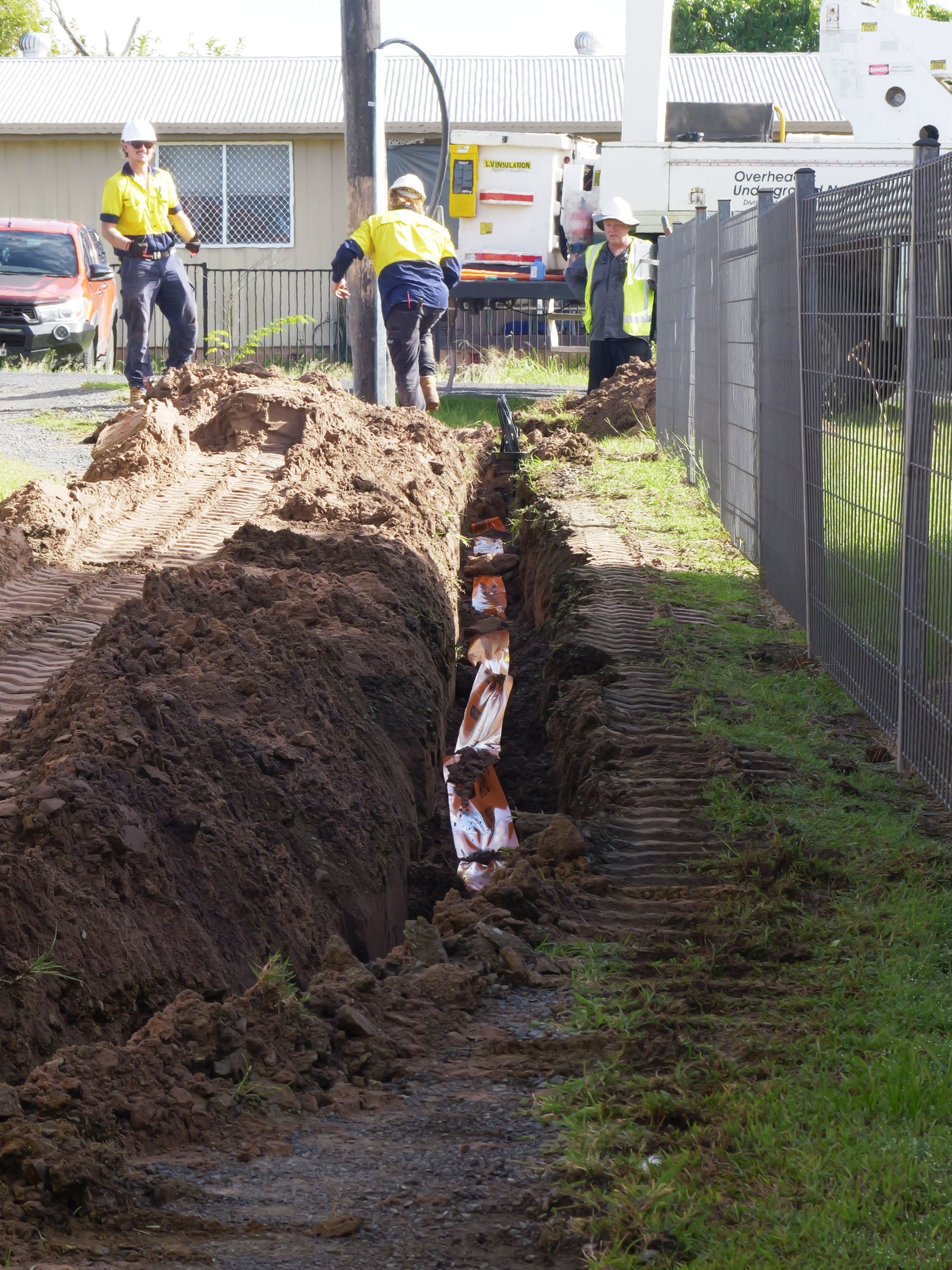 Construction workers digging a trench; orange pipes in trench, near a fence and utility pole— Rob Ward Electrical Services in West Ballina, NSW