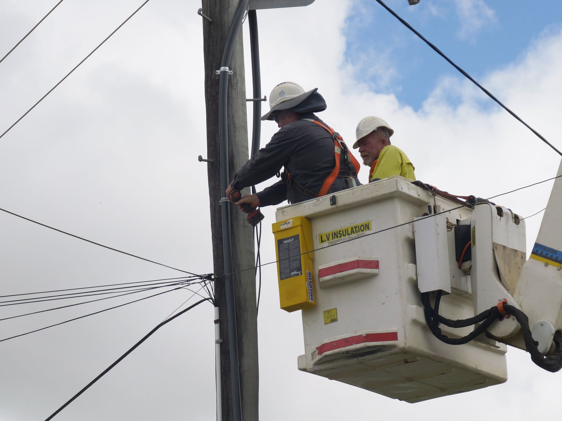 Two utility workers in a bucket truck working on power lines attached to a gray pole against a cloudy sky— Rob Ward Electrical Services in West Ballina, NSW