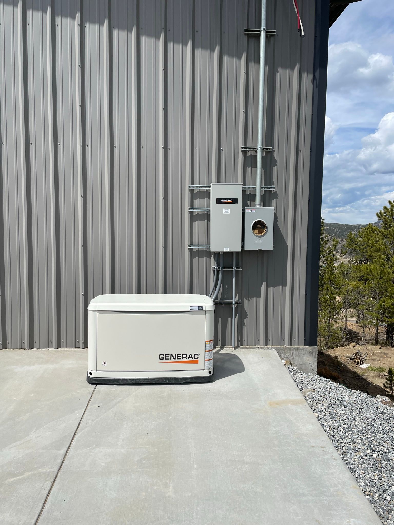 Generator and electrical panel on a gray metal building exterior. Concrete and gravel surround. Cloudy sky.