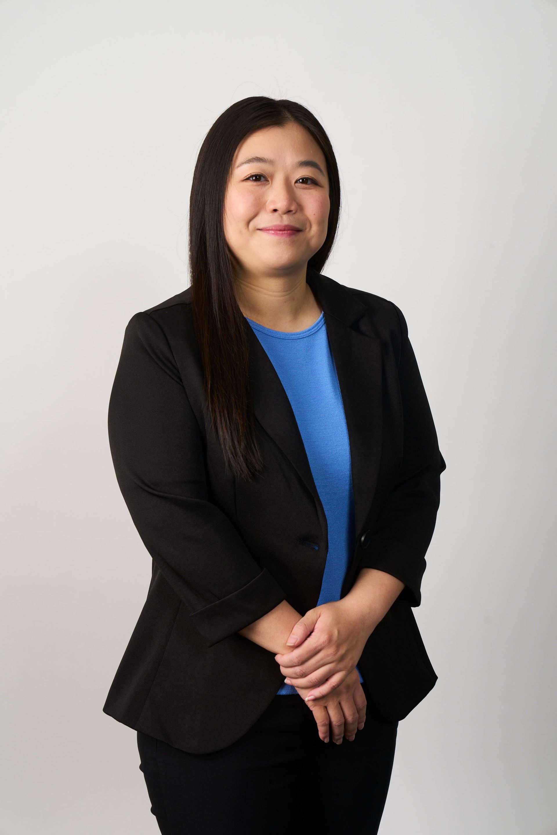 A person with long dark hair wearing a black blazer over a bright blue shirt, posing against a white background
