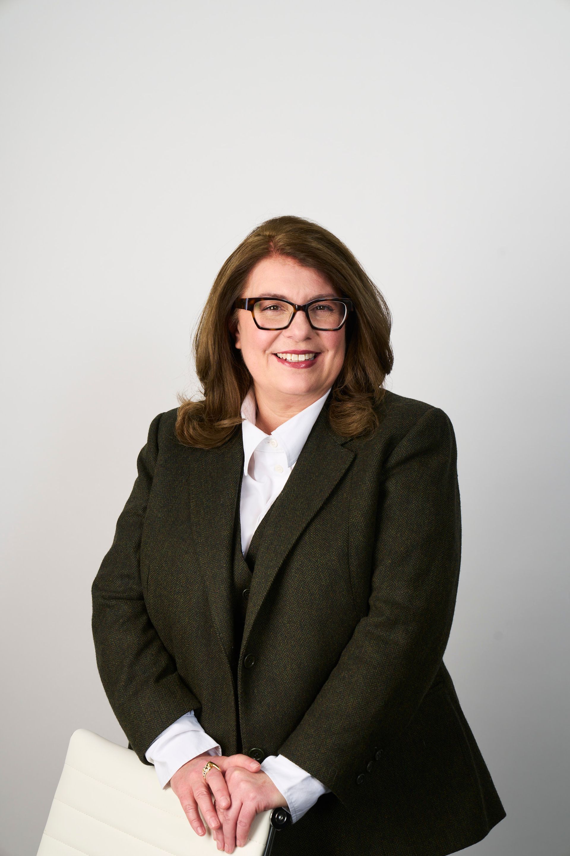 A person with shoulder-length brown hair and glasses, wearing a dark textured blazer and white shirt, smiling