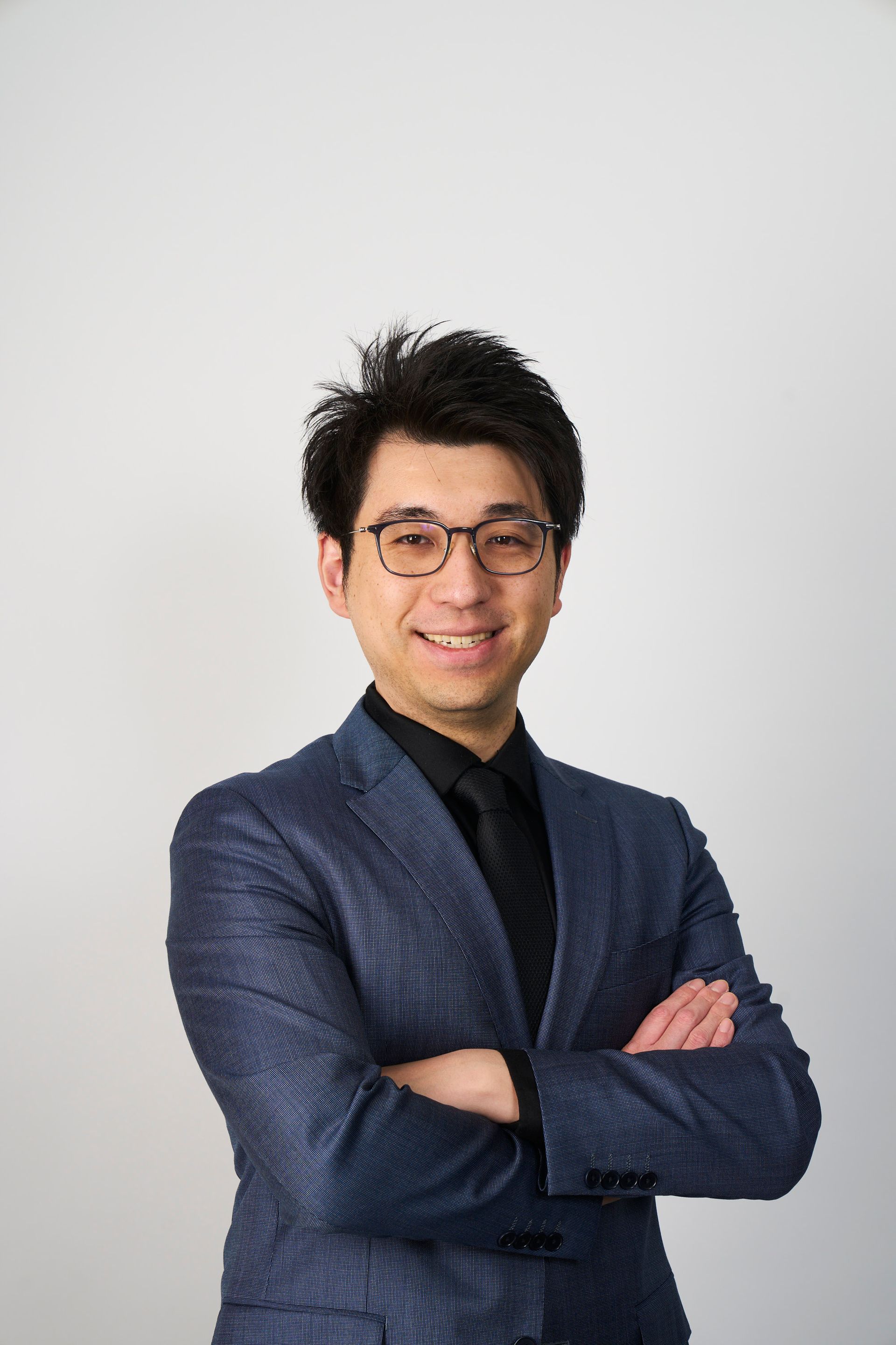 A professional portrait of a person in a blue textured blazer and glasses, smiling against a grey wall.