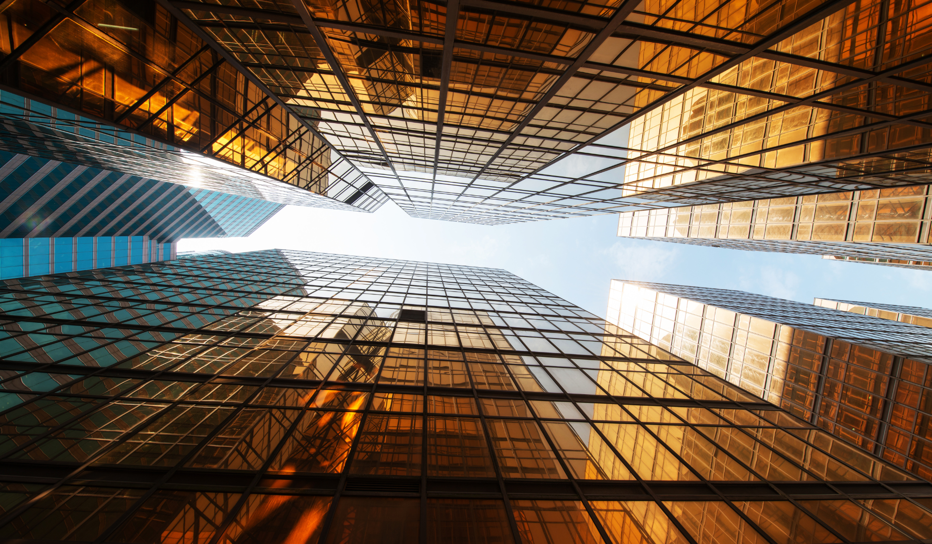 A low-angle view looking up at modern glass skyscrapers with warm golden reflections and a strip of bright blue sky.
