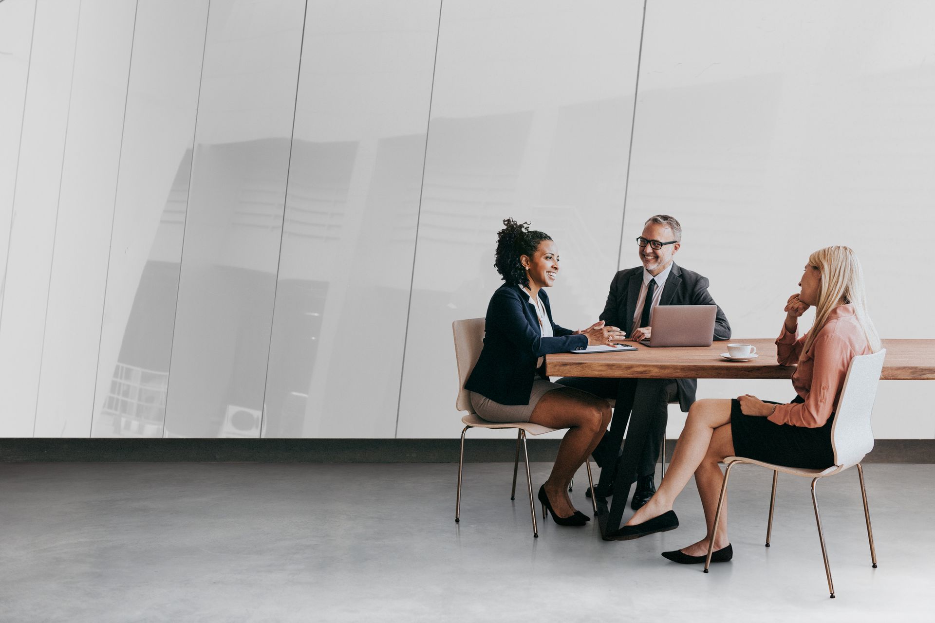 Three people in business attire sit at a conference table in a modern, white office space, engaged in a discussion