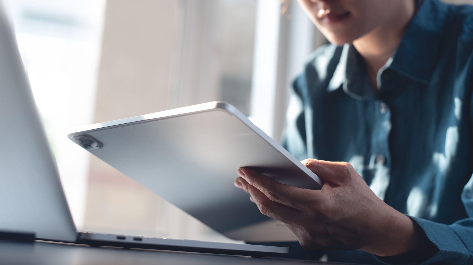 A person in a dark shirt sitting at a desk, looking at a tablet held in their hands next to an open laptop