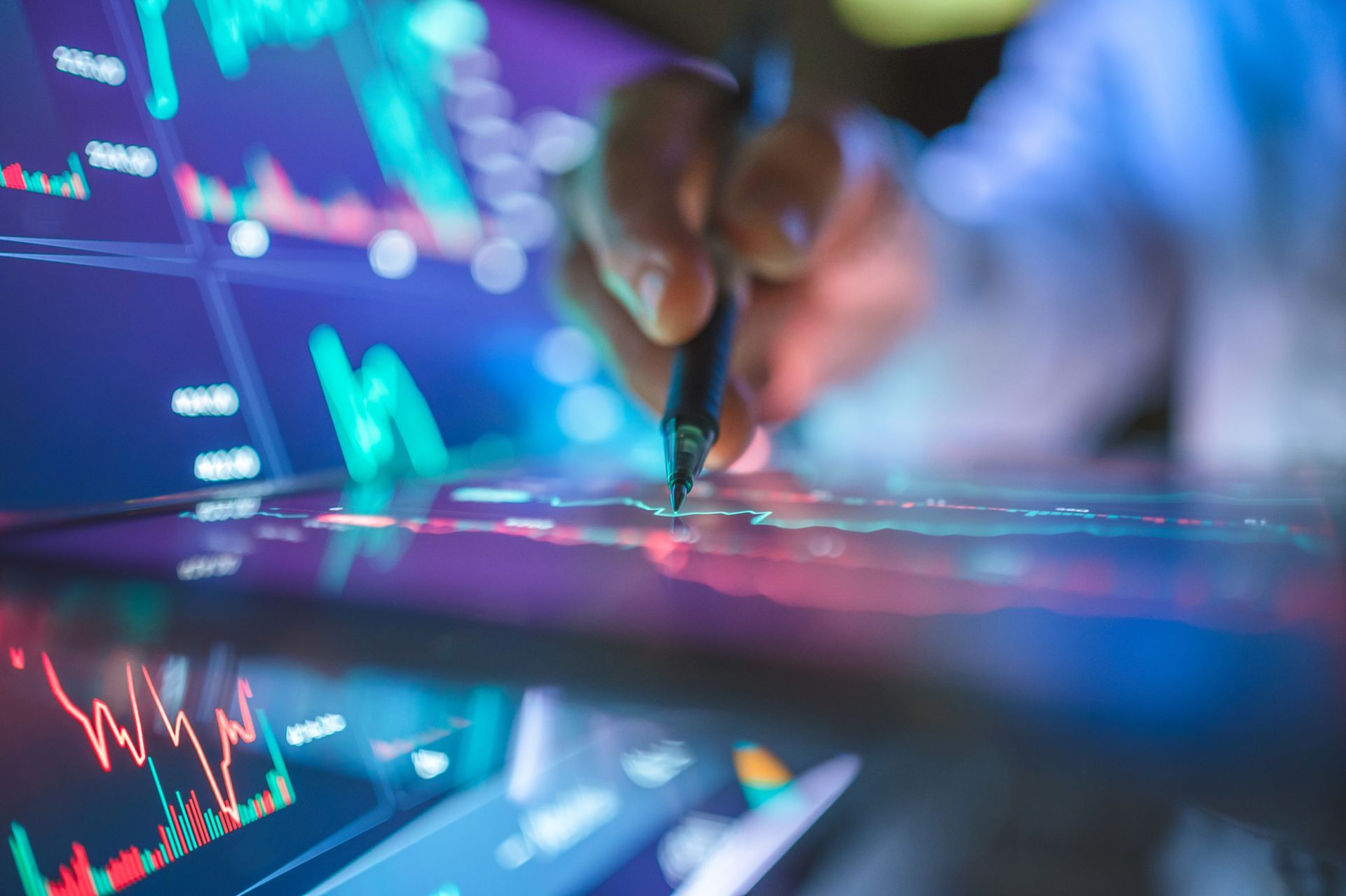 A hand uses a pen on a digital screen displaying glowing financial stock market charts in a dark