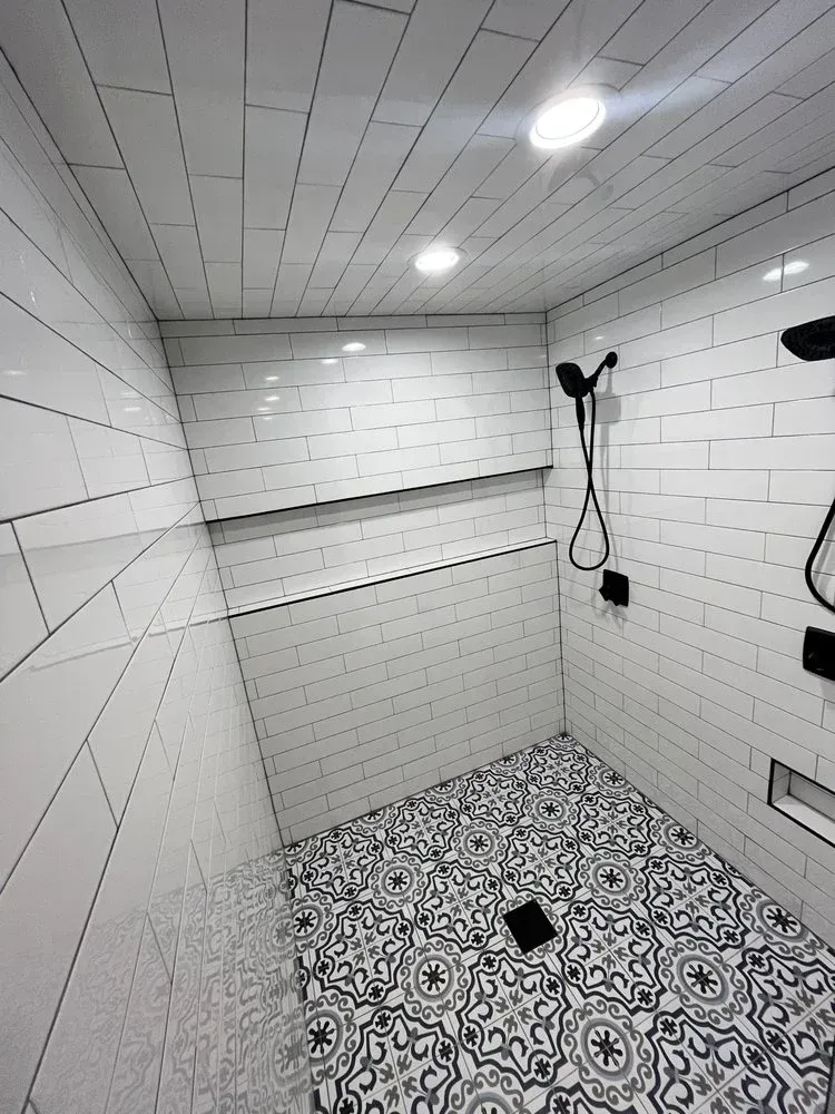 Modern white-tiled shower stall with handheld shower, overhead light, and patterned floor tiles.